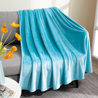 Plaid bleu en polaire étendu sur un canapé gris avec un bouquet de fleurs oranges dans un vase posé sur un bout de guéridon à coté.