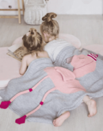deux petites filles allongées sur le sol de dos en train de lire un livre avec un plaid flamand rose sur elles