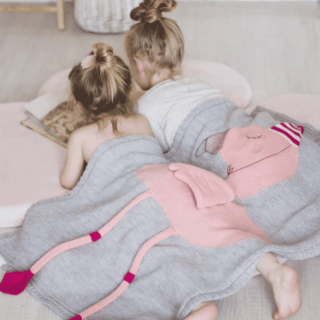 deux petites filles allongées sur le sol de dos en train de lire un livre avec un plaid flamand rose sur elles
