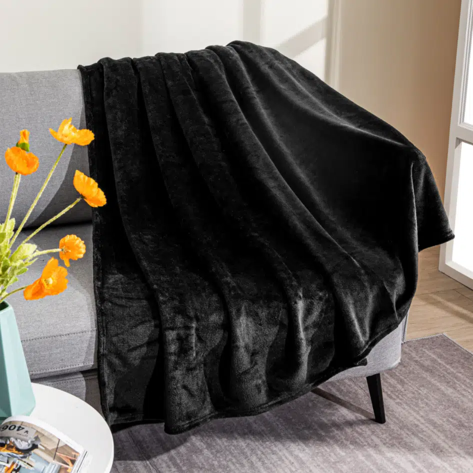 Plaid noir en polaire étendu sur un canapé gris avec un bouquet de fleurs orangées dans un vase à coté posé sur un bout de guéridon.