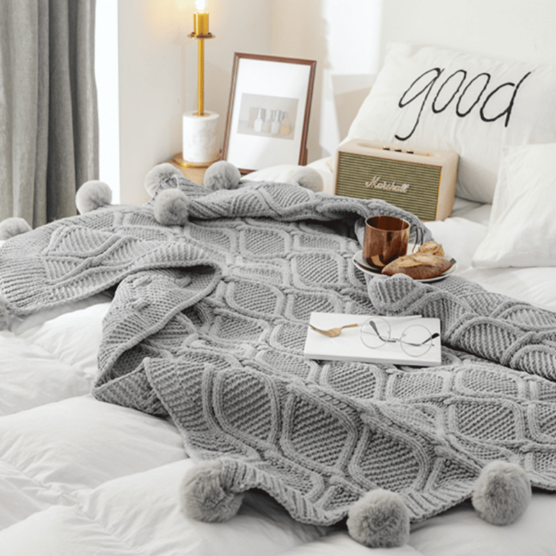 Plaid gris tricoté avec pompons dans une chambre sur un lit blanc avec un livre, une assiette de nourriture et des lunettes.