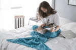 Femme brune sur un lit blanc en train de lire un magazine dans un plaid sirène tricoté en bleu