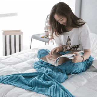 Femme brune sur un lit blanc en train de lire un magazine dans un plaid sirène tricoté en bleu