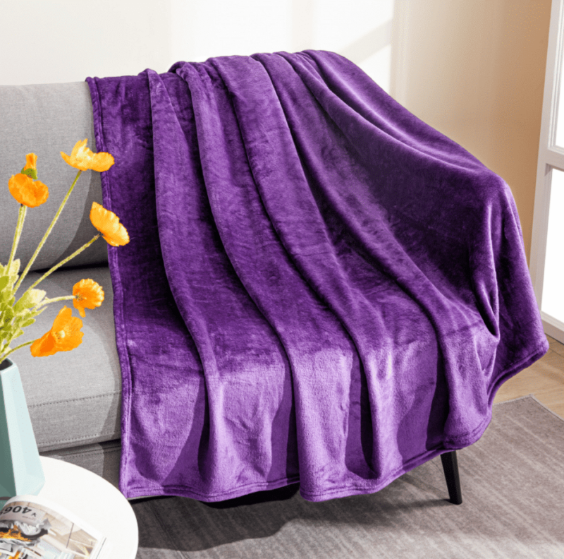 Plaid en polaire violet étendu sur un canapé gris à coté d'un bouquet de fleurs orangées dans un vase posé sur un bout de guéridon.
