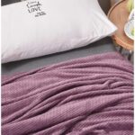 Plaid rose violet polaire effet piqué sur un lit près d'un coussin rectangulaire blanc avec petites inscriptions noires