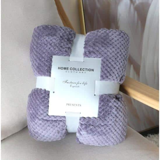 Posé sur un fauteuil gris, un plaid violet lilas finition piqué en polaire, plié et entouré d'un ruban blanc avec une étiquette blanche carré au centre.