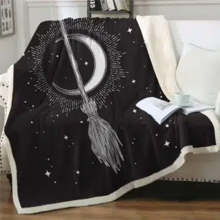 Un plaid en polaire au fond noir avec des dessins blancs :un quartier de lune qui scintille, des étoiles et un balai par-dessus. Le plaid est étendu sur un canapé blanc avec un livre ouvert posé dessus à côté d'un coussin blanc.