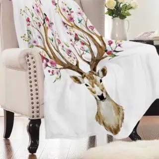 On voit un joli fauteuil crapaud beige avec des clous en laiton et des pieds en bois sombre. Dessus, est posé un plaid blanc avec un grand cerf dont les bois sont fleuris de fleurs roses.