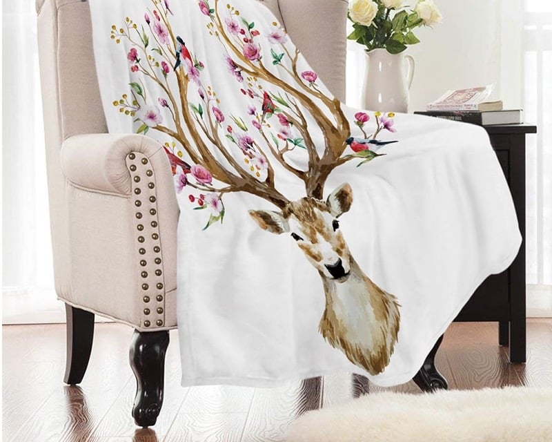 On voit un joli fauteuil crapaud beige avec des clous en laiton et des pieds en bois sombre. Dessus, est posé un plaid blanc avec un grand cerf dont les bois sont fleuris de fleurs roses.