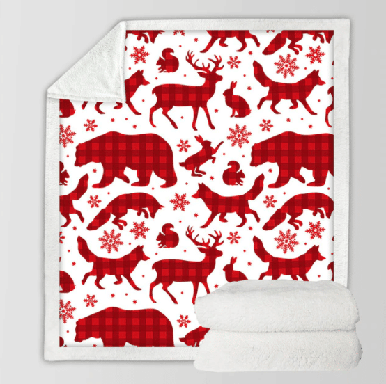 Plaid polaire blanc avec des formes d'animaux de la forêt comme des rennes, des ours, renards , lapins en imprimé rouge vichy et des flocons de neige. Il y a deux plaids blancs en polaire pliés l'un sur l'autre en bas à droite de l'image .