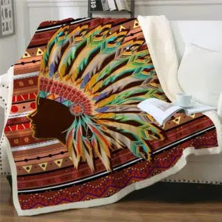 Plaid coloré dans des tons bruns, rouges vert et orangé avec une tête d'indien avec une coiffe étendu sur un canapé blanc. Il y a un livre avec une tasse dessus.