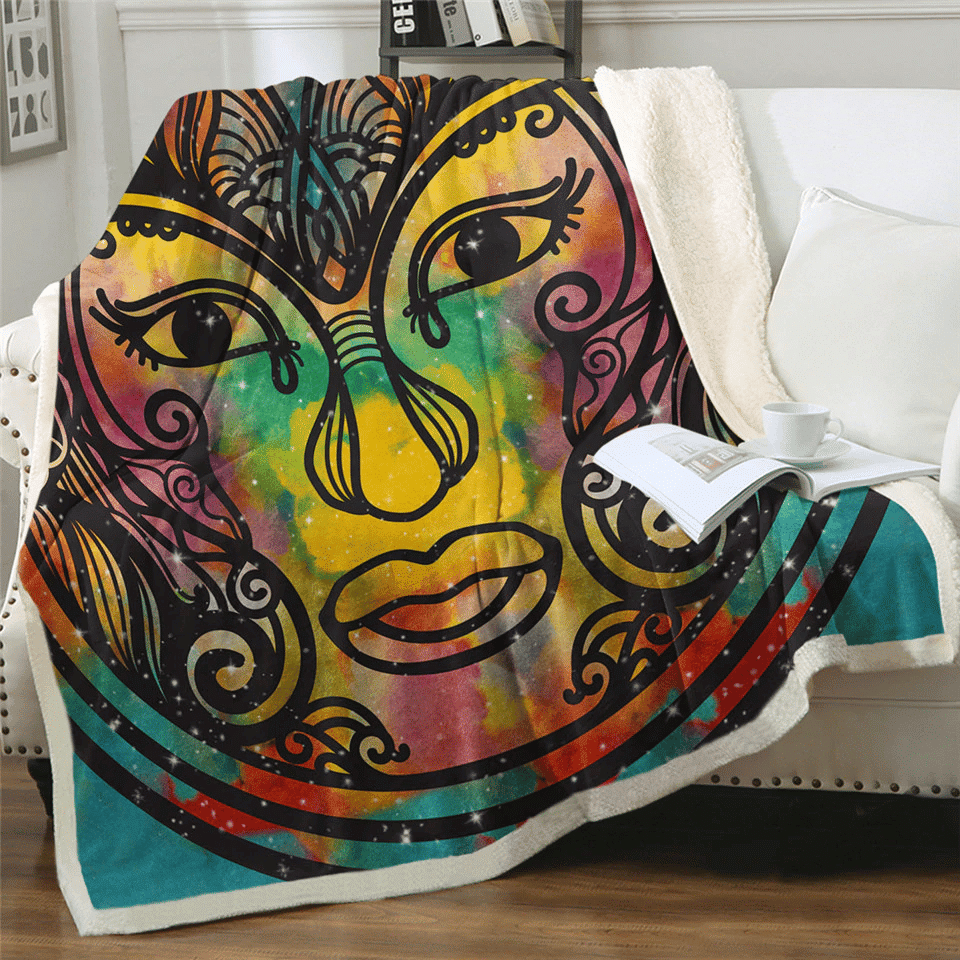 Plaid polaire multicolore imprimé à motif visage ethnique sur un canapé blanc. Il y a un livre ouvert dessu avec une tasse blanche par dessus et un coussin blanc à côté.