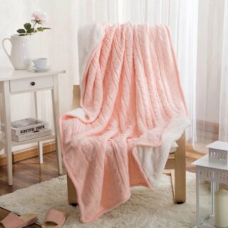 Plaid rose en maille torsadée étendu sur un fauteuil avec pieds en bois sur un tapis beige à coté d'une petite console blanche avec une cruche posé dessus et une rose dedans.