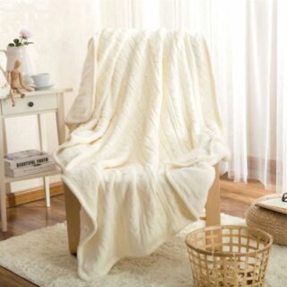 Plaid blanc en maille torsadée étendu sur un fauteuil avec pieds en bois sur un tapis en coton blanc avec un panier tressé ajouré à coté.