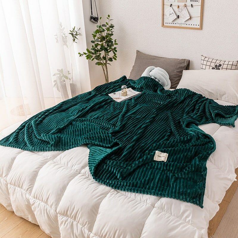 Plaid en flanelle velours vert étendu sur un lit sur une couette matelassée blanche dans une chambre au décor épuré avec rideaux légers blancs transparent et un olivier près du lit .