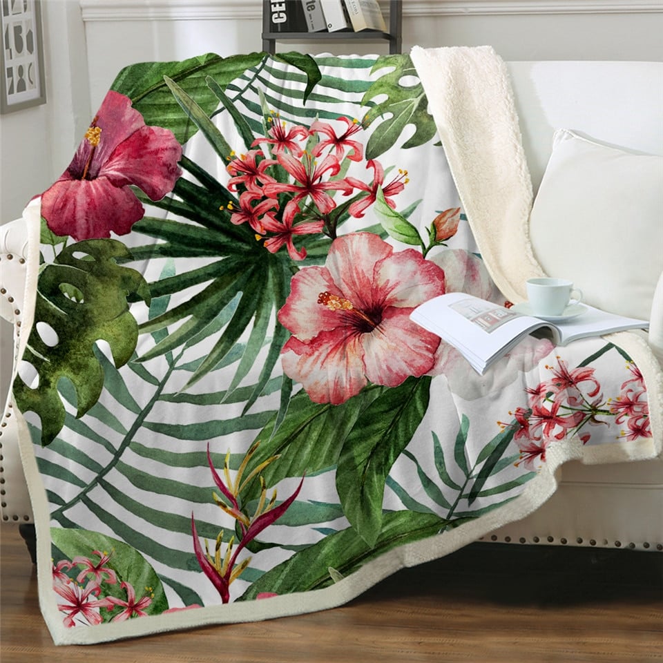 Plaid polaire avec hibiscus et plantes tropical étendu sur un canapé blanc à côté d'un coussin blanc et d'un livre ouvert avec une tasse posée dessus.