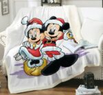Plaid blanc avec Mickey et Minnie avec des habits d'hiver rouges bordés de fourrure. Le plaid est étendu sur un canapé blanc, un livre ouvert avec une tasse posée sur le dessus à côté d'un coussin blanc