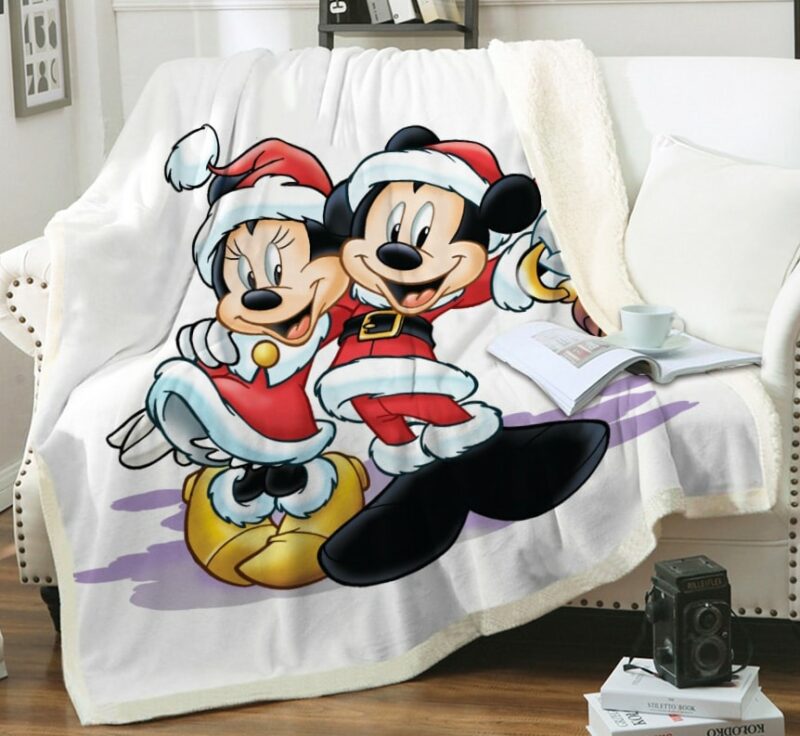 Plaid blanc avec Mickey et Minnie avec des habits d'hiver rouges bordés de fourrure. Le plaid est étendu sur un canapé blanc, un livre ouvert avec une tasse posée sur le dessus à côté d'un coussin blanc