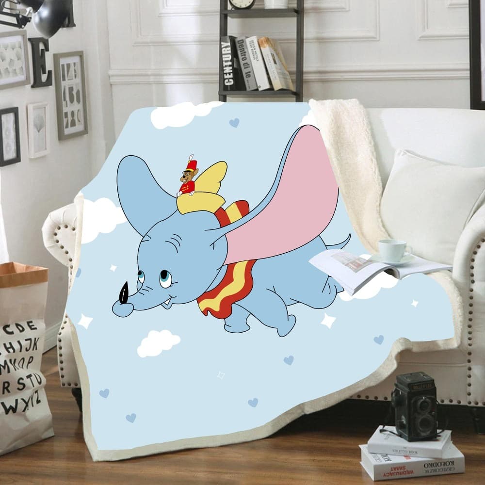Plaid bleu clair avec Dumbo qui vole dans le ciel avec son ami la souris sur la tête. Le plaid est étendu sur un canapé blanc avec un livre et une tasse posée dessus à coté d'un coussin blanc. Il y a deux livres posés sur le sol en parquet avec un appareil dessus.