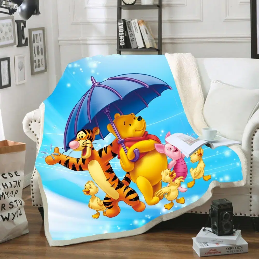 Plaid coloré avec Winnie l'ourson, Tigrou et Porcinet avec un parapluie bleu foncé sur fond bleu ciel. Il est étendu sur un canapé blanc dans un décor de salon avec une étagère en arrière-plan et des cadres sur les murs.