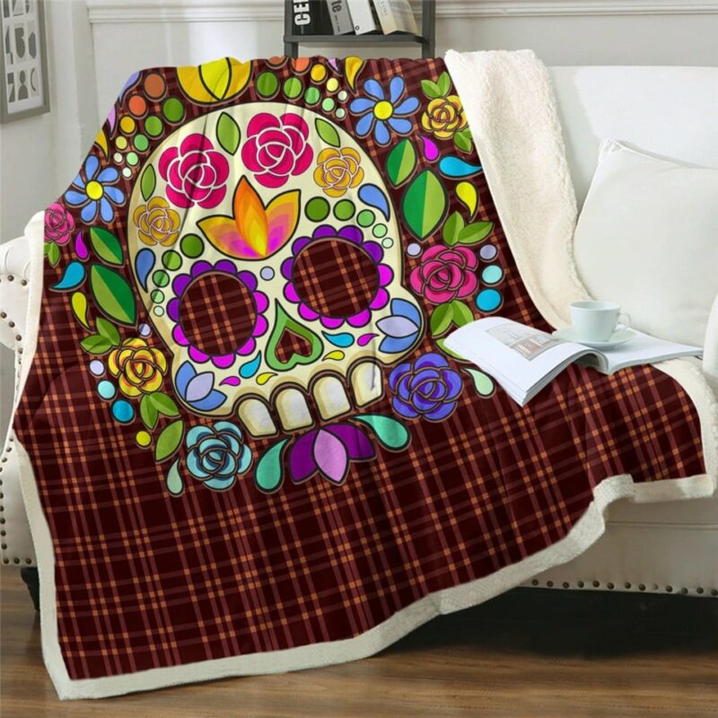 Plaid polaire avec crâne de tête mexicaine avec fleurs colorées et imprimé tissu écossais marron, étendu sur un canapé blanc et débordant sur le sol en parquet marron. Il y a un livre ouvert avec une tasse blanche posée dessus.