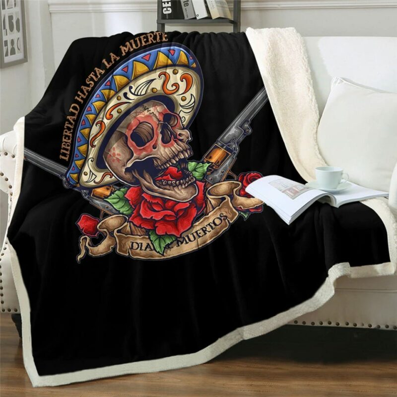 Plaid polaire noire avec tête de mort mexicaine colorée avec un sombrero, des roses rouges et deux pistolets. Il est étendu sur un canapé blanc et déborde sur le sol en parquet marron. Il y a un livre ouvert posé dessus avec une tasse blanche par-dessus.