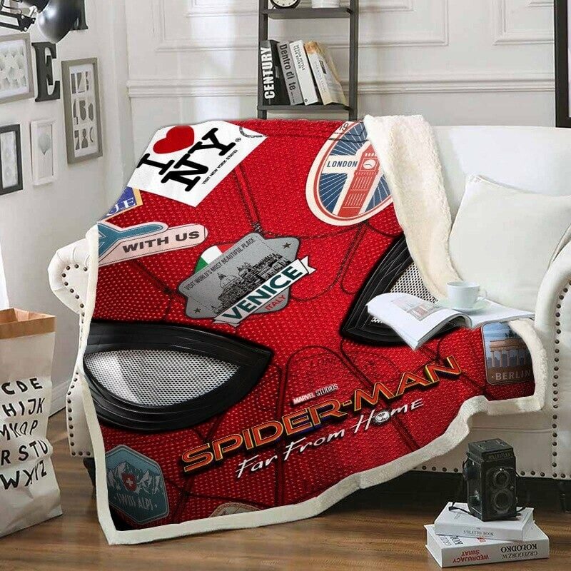 Plaid polaire rouge bleu et noir Spiderman au look rétro avec les yeux noir et un patchwork d'écussons de villes. Ce plaid est étendu sur un canapé blanc dans un décor de salon avec un livre et une tasse posée dessus .