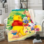 Plaid polaire coloré avec Winnie l'ourson, Tigrou et la souris cueillant des fleurs. Il est étendu sur un canapé blanc à coté d'un coussin avec un livre ouvert et une tasse posée dessus dans un décor de salon.
