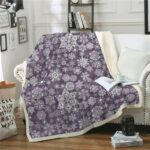 Plaid polaire mauve avec flocons de neige en rosaces blancs, étendu sur un canapé blanc à côté d'un coussin blanc. Il y a un livre ouvert avec une tasse dessus posée. Au sol , deux livres sont posés avec un appareil noir dessus.