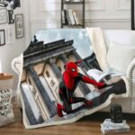 Plaid polaire avec Spiderman en mouvement au sol dans la rue avec un grand bâtiment historique en arrière plan. Ce plaid est étendu sur un canapé blanc dans un décor de salon avec un livre et une tasse posée dessus.