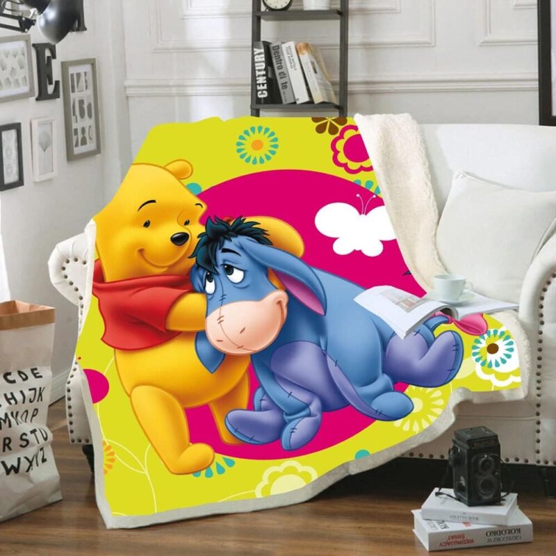 Plaid polaire coloré au fond jaune dominant avec Winnie l'ourson qui cajole son ami Bourriquet. Il est étendu sur un canapé blanc à coté d'un coussin dans un décor de salon avec une étagère en arrière-plan et des cadres sur le mur .