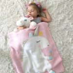 une petite fille dormant sur un tapis doux blanc avec une peluche licorne et un plaid rose avec une licorne blanche