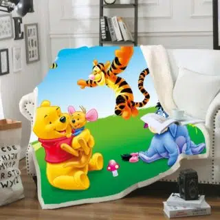Plaid polaire coloré avec personnages Winnie l'ourson, Tigrou, Bourriquet sur l'herbe. Il est étendu sur un canapé blanc avec un coussin à coté et un livre ouvert dessus dans un décor de salon.