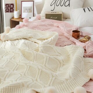 Plaids tricotés beige et rose avec motifs losanges et pompons étendu sur un lit dans une ambiance cocooning. Il y a des coussins blancs sur le lit une enceinte, une tasse et des viennoiseries.