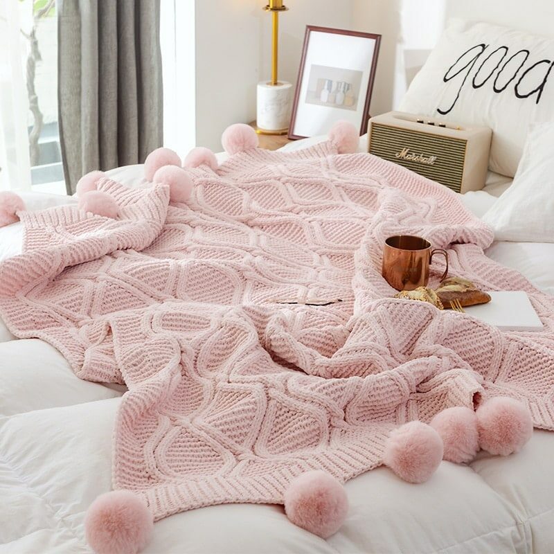 Plaid rose tricoté avec motifs losanges et pompons étendu sur une couette blanche matelassée sur un lit dans une ambiance cocooning. Il y une enceinte, une tasse cuivré et des viennoiseries posés dessus.