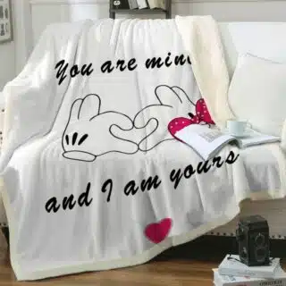 Plaid blanc avec gant de Minnie et de Mickey se joignant pour faire un cœur avec un message d'amour "You are mine and I am yours ".Il est étendu sur un canapé blanc avec un livre ouvert posé dessus avec une tasse à côté d'un coussin blanc.