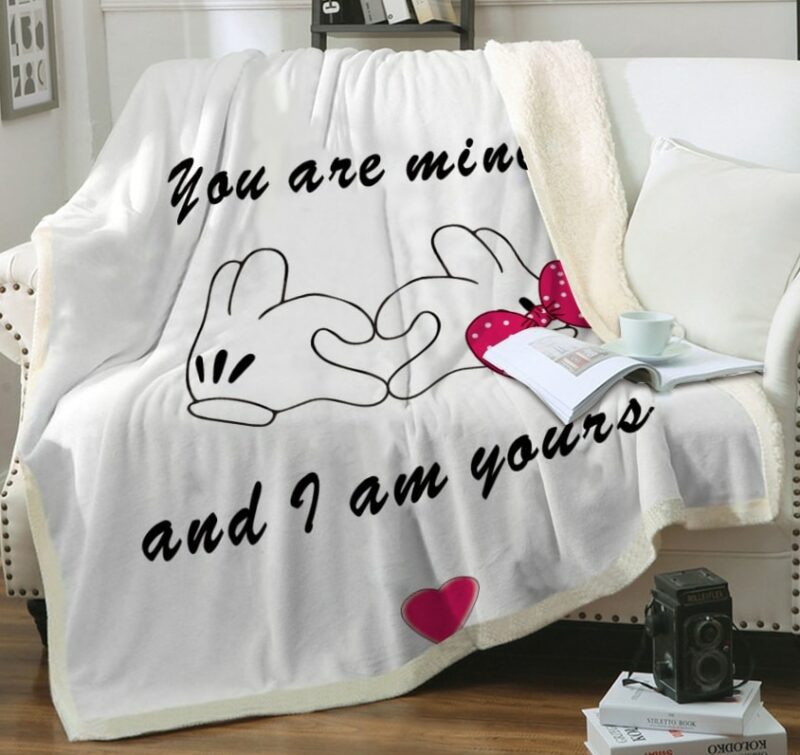 Plaid blanc avec gant de Minnie et de Mickey se joignant pour faire un cœur avec un message d'amour "You are mine and I am yours ".Il est étendu sur un canapé blanc avec un livre ouvert posé dessus avec une tasse à côté d'un coussin blanc.