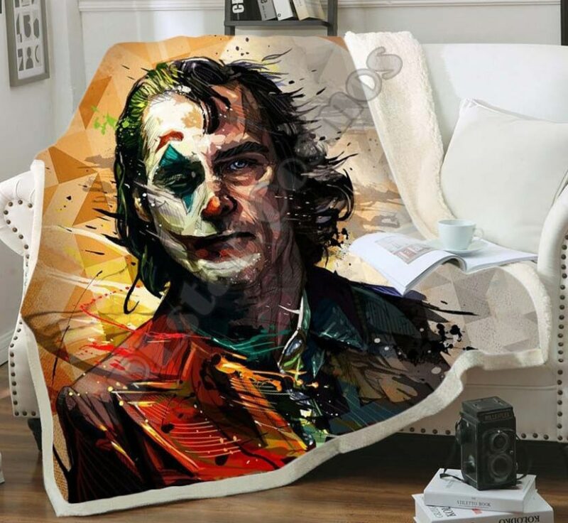Plaid polaire multicolore abstrait double face de Joker avec la moitié du visage avec maquillage de clown et l'autre sans. Il est étendu sur un canapé blanc et déborde sur le parquet marron au sol.
