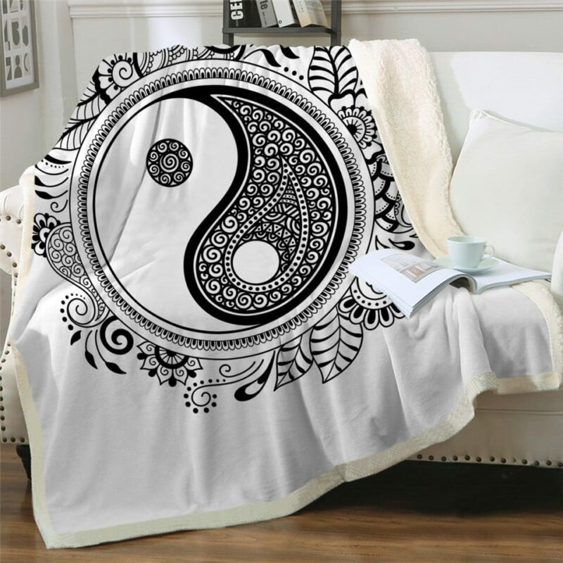 Plaid polaire noir et blanc avec motif Yin et Yang avec des fleurs autour. Il est posé sur un canapé blanc avec un livre ouvert avec tasse posée dessus à côté d'un coussin blanc.