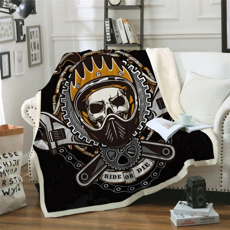 Plaid polaire noir et orange avec tête de mort dans un casque de motard entouré de chaines et d'outils avec le message "Ride or dye". Il est étendu sur un canapé blanc et déborde sur le sol en parquet marron dans un décor de salon. Il y a un coussin blanc à côté avec un livre ouvert dessus et une tasse blanche posée par dessus.
