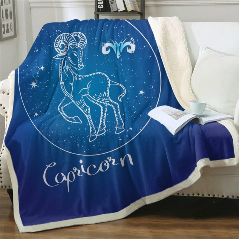 Plaid polaire bleu nuit signe astrologique capricorne sur un canapé blanc. Il y a un livre ouvert avec une tasse blanche par dessus.