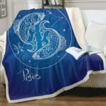 Plaid bleu nuit signe astrologique poisson sur un canapé blanc avec un livre ouvert posé dessus et une tasse blanche par dessus.