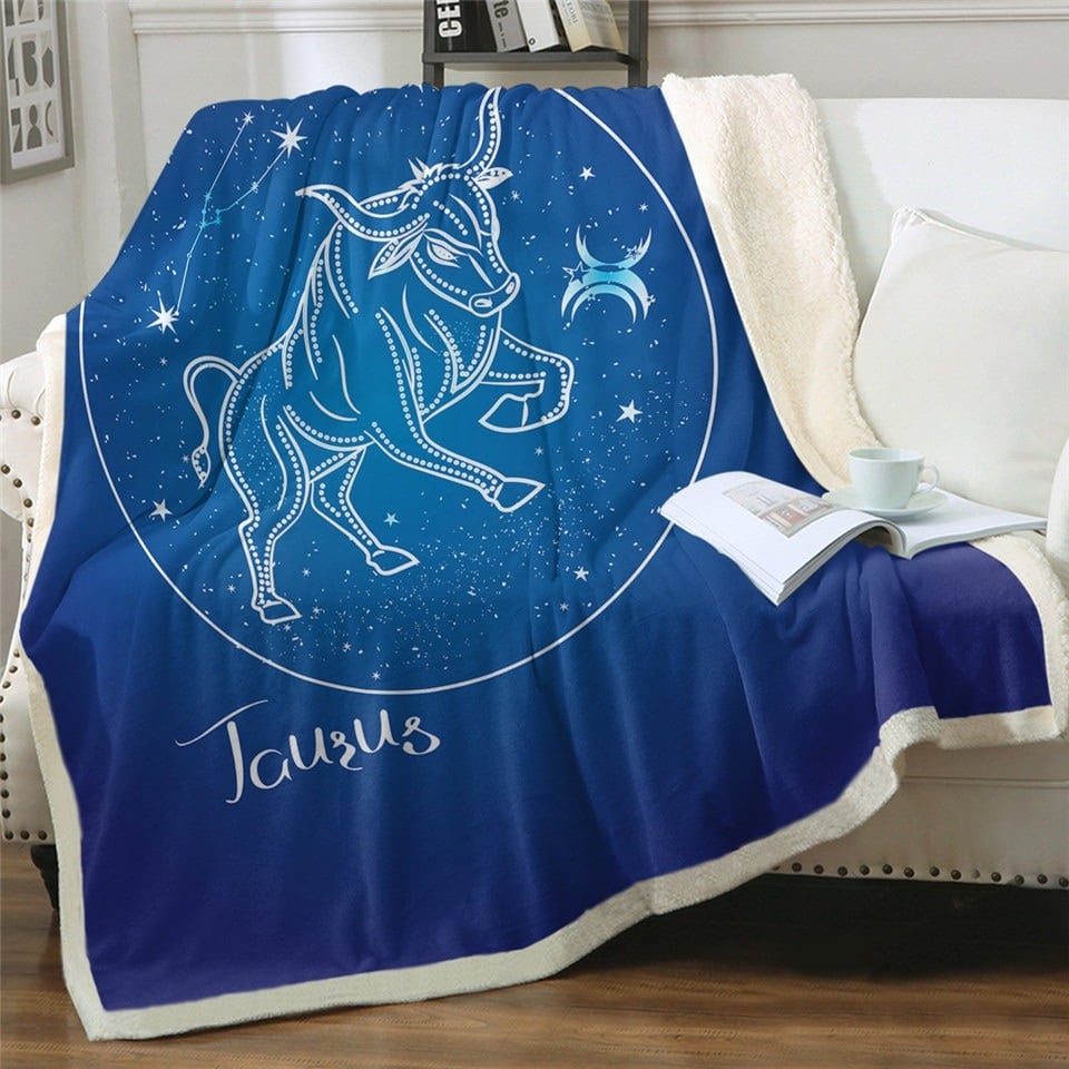 Plaid polaire bleu nuit signe astrologique taureau sur un canapé blanc. Il y a un livre ouvert posé dessus avec une tasse blanche par-dessus.