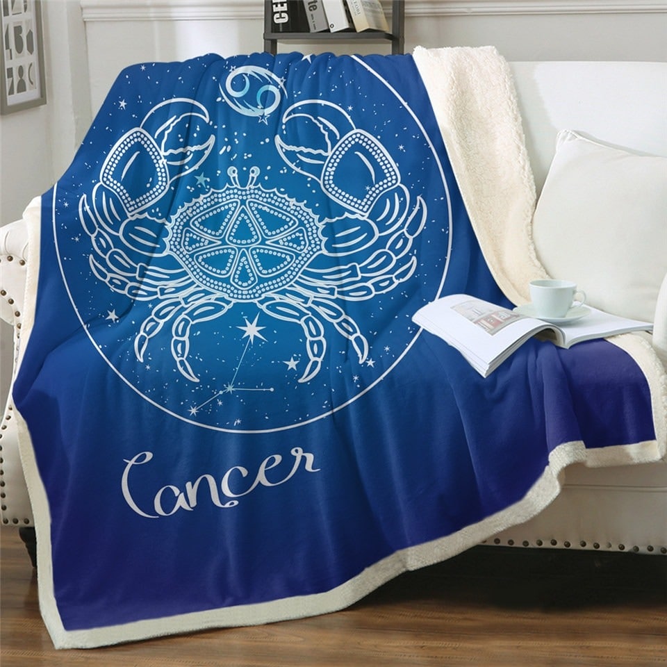 Plaid polaire bleu nuit signe astrologique cancer sur un canapé blanc. Il ya un livre ouvert dessus avec une tasse blanche posée par-dessus.