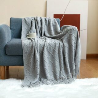 Plaid tricoté avec losanges en relief et franges étendu sur un fauteuil bleu et débordant sur un tapis en fourrure blanche. Il y a une tasse blanche posée dessus