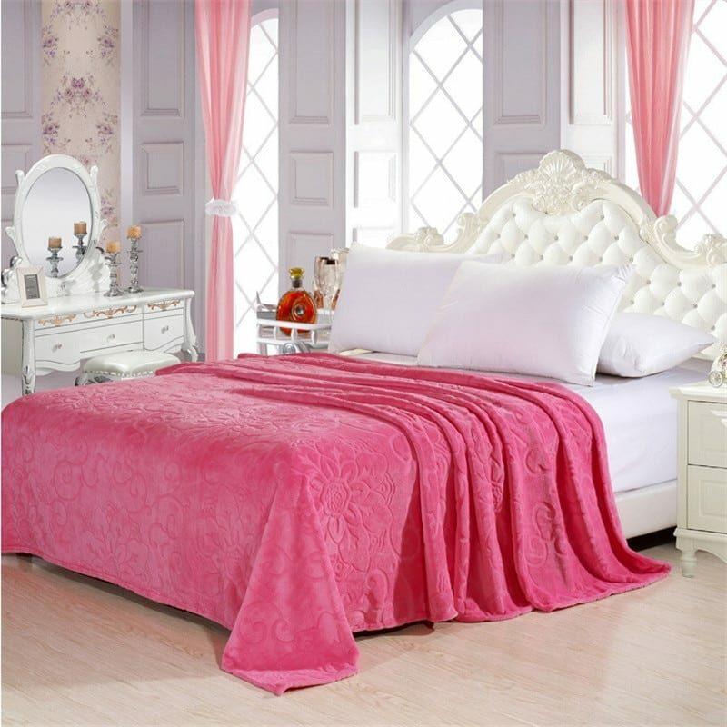 Couverture en velours rose avec motifs en reliefs fleuris, étendue sur un lit 2 places avec deux coussins blancs et une tête de lit blanche capitonnée. La chambre a un décor baroque avec une coiffeuse blanche et des grandes fenêtres et des rideaux roses derrière le lit.