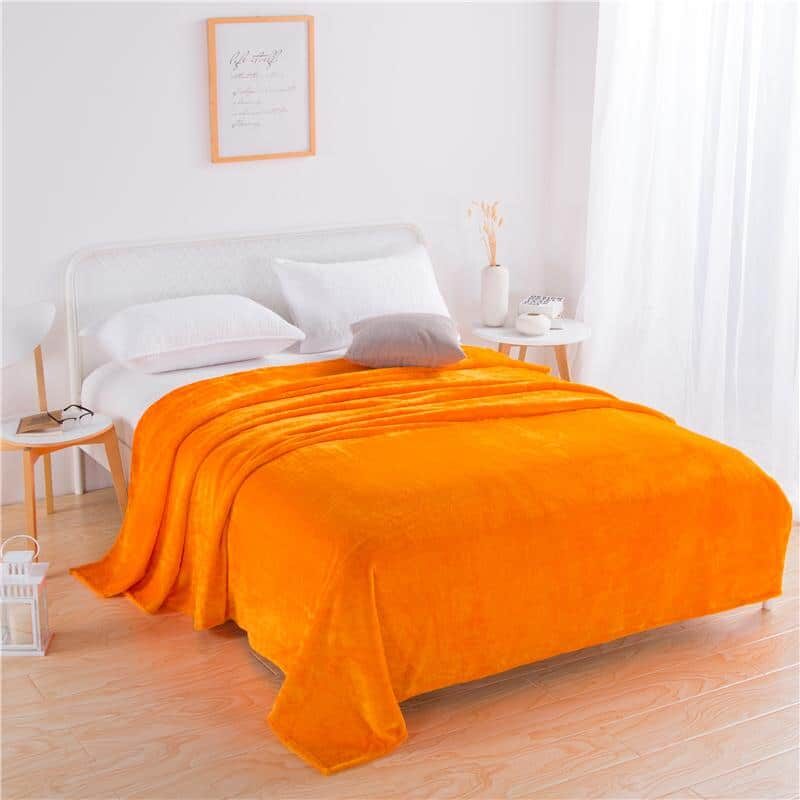 Grande couverture orange en flanelle couvrant entièrement un lit deux places sur une literie blanche dans une chambre épuré. On y voit deux petites et simples tables de nuit avec des lampes blanches et un cadre beige au -dessus d'une tête de lit gris clair. Le sol est en parquet beige clair.
