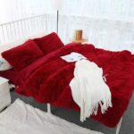 Dans un décor de chambre blanc épuré, un grand couvre-lit rouge en fausse fourrure recouvrant totalement un lit 2 places avec des coussins rectangulaires assortis. Il y a un grand châle blanc avec un magazine posé dessus.