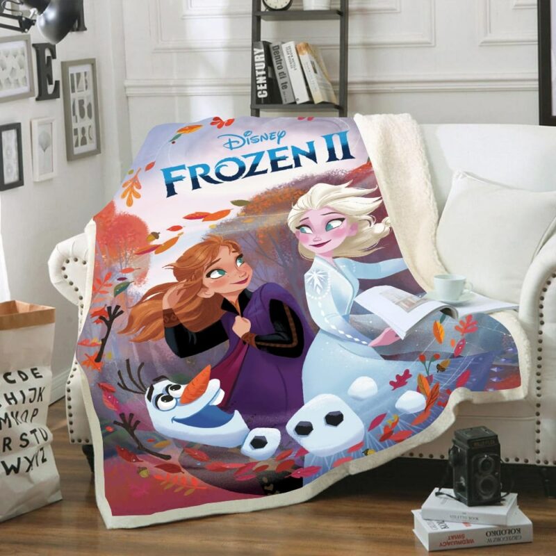 Plaid polaire avec "Frozen II" écrit dessus et les personnages de Elsa, Anna et Olaf en imprimé coloré. Le plaid est étendu sur un canapé blanc dans un décor de salon avec parquet en bois, cadres sur le mur et étagère rempli d'objets en arrière-plan.