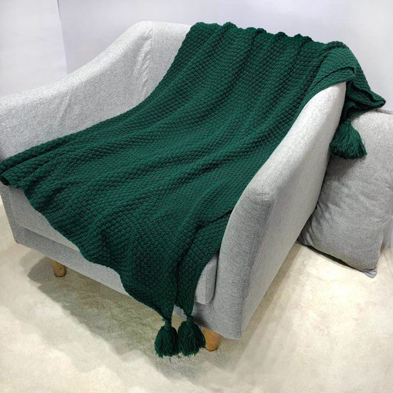 Plaid vert tricoté avec pompons sur les coins étendu sur un fauteuil gris avec coussin gris sur un sol en moquette beige